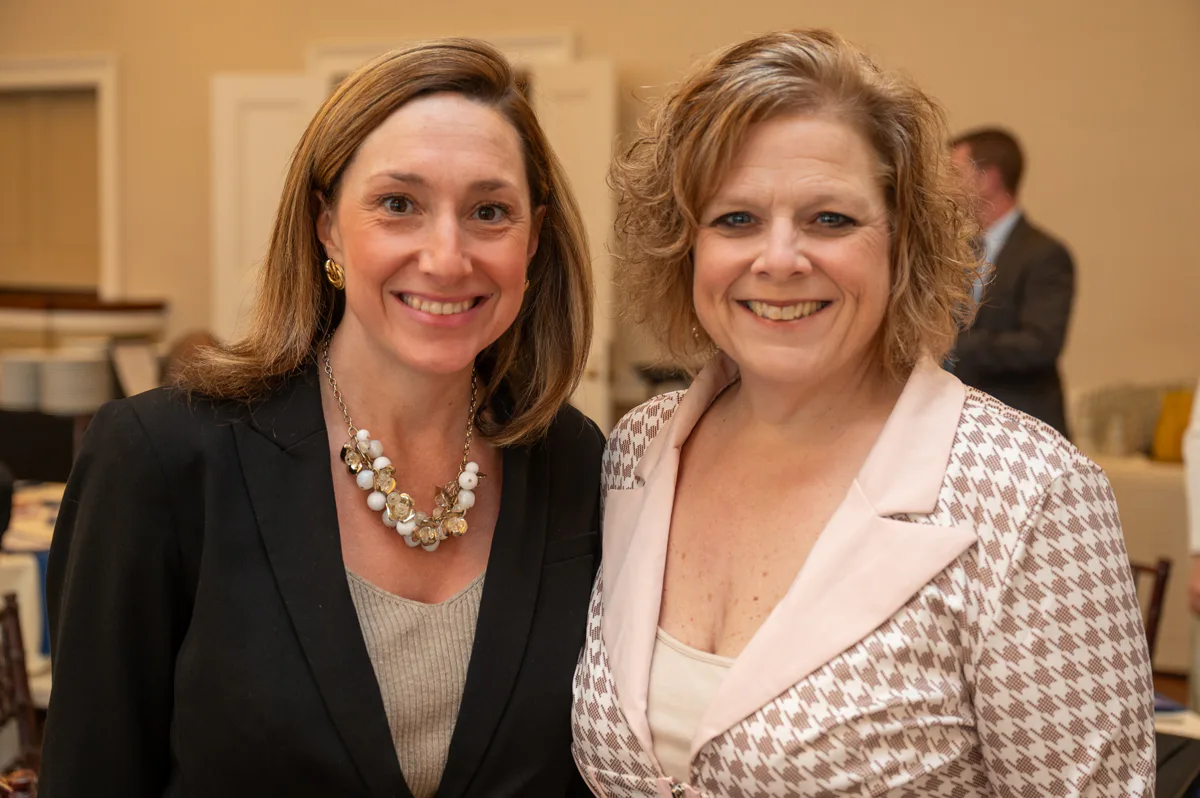Two women smile side by side at an event, one in a black top with pearls