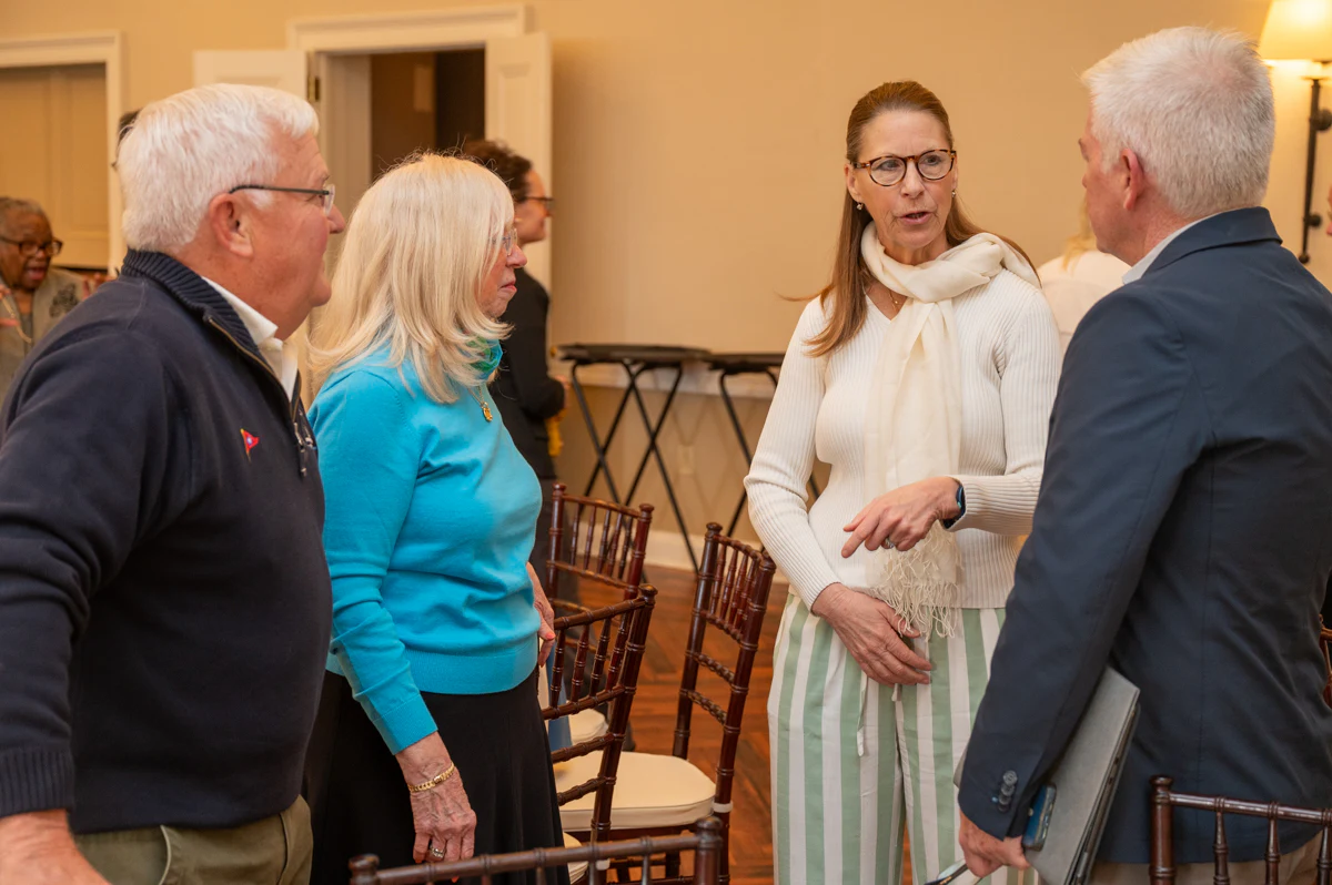 Four people chat in a room, with a woman in a white blouse gesturing