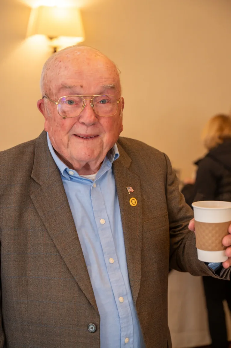 Older smiling man in gray blazer and blue shirt holds a coffee cup