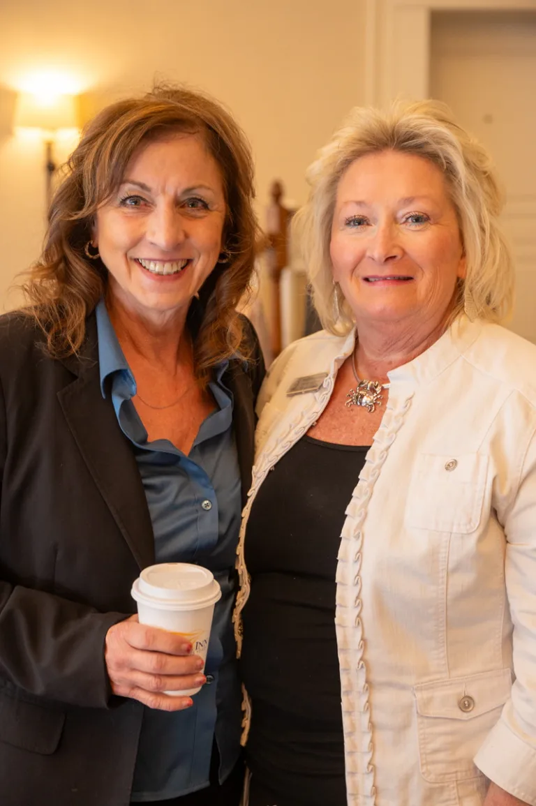 Two smiling women stand together, one holding a coffee cup