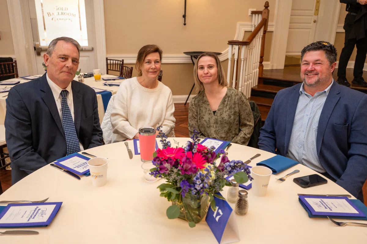 Four people sit at a banquet table with flowers and blue program folders