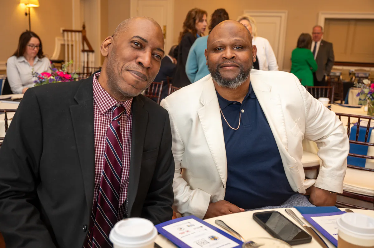 Two men sit at a table with blue program folders, one in a white blazer