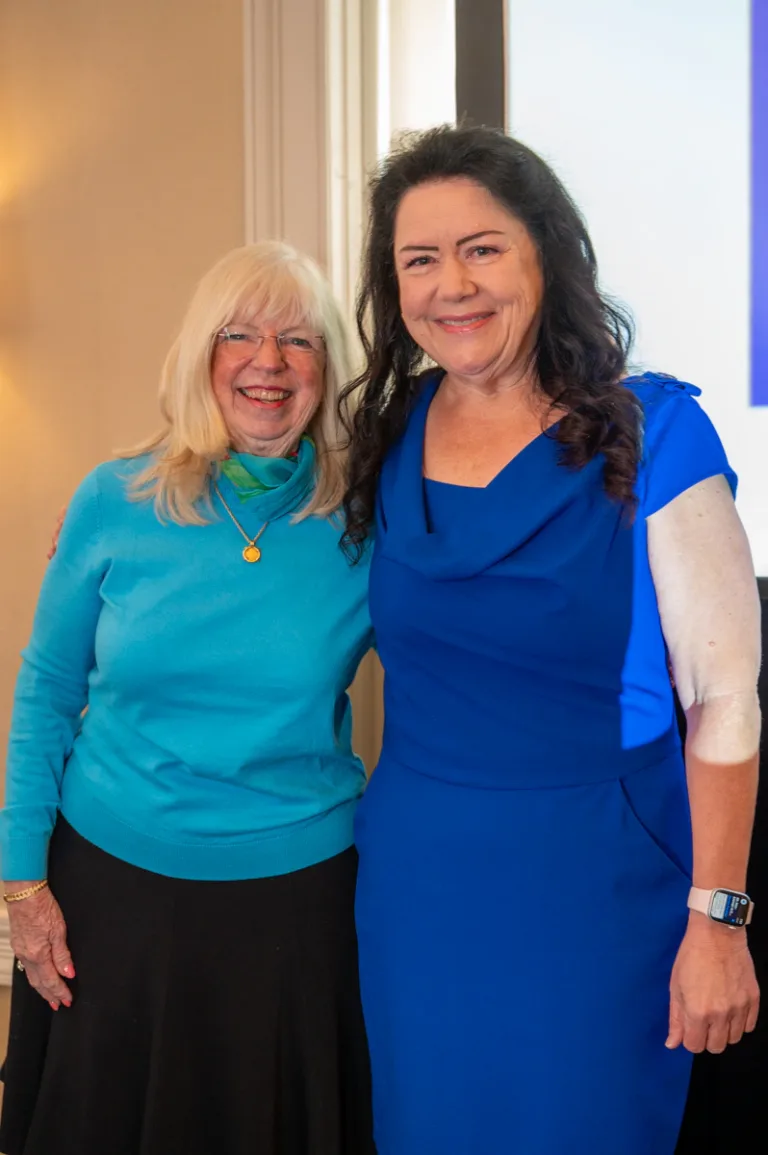 Two women smile together, one in turquoise sweater and one in blue dress