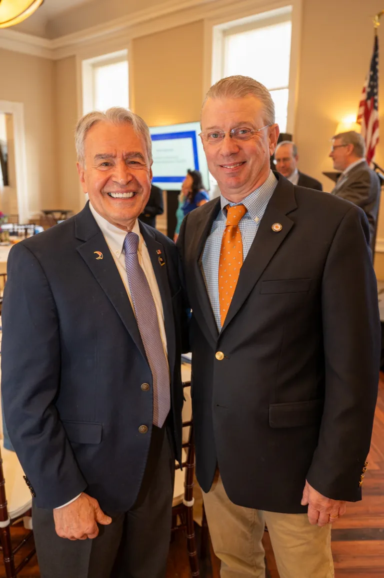 Two men in dark suits smile together, one wearing an orange tie