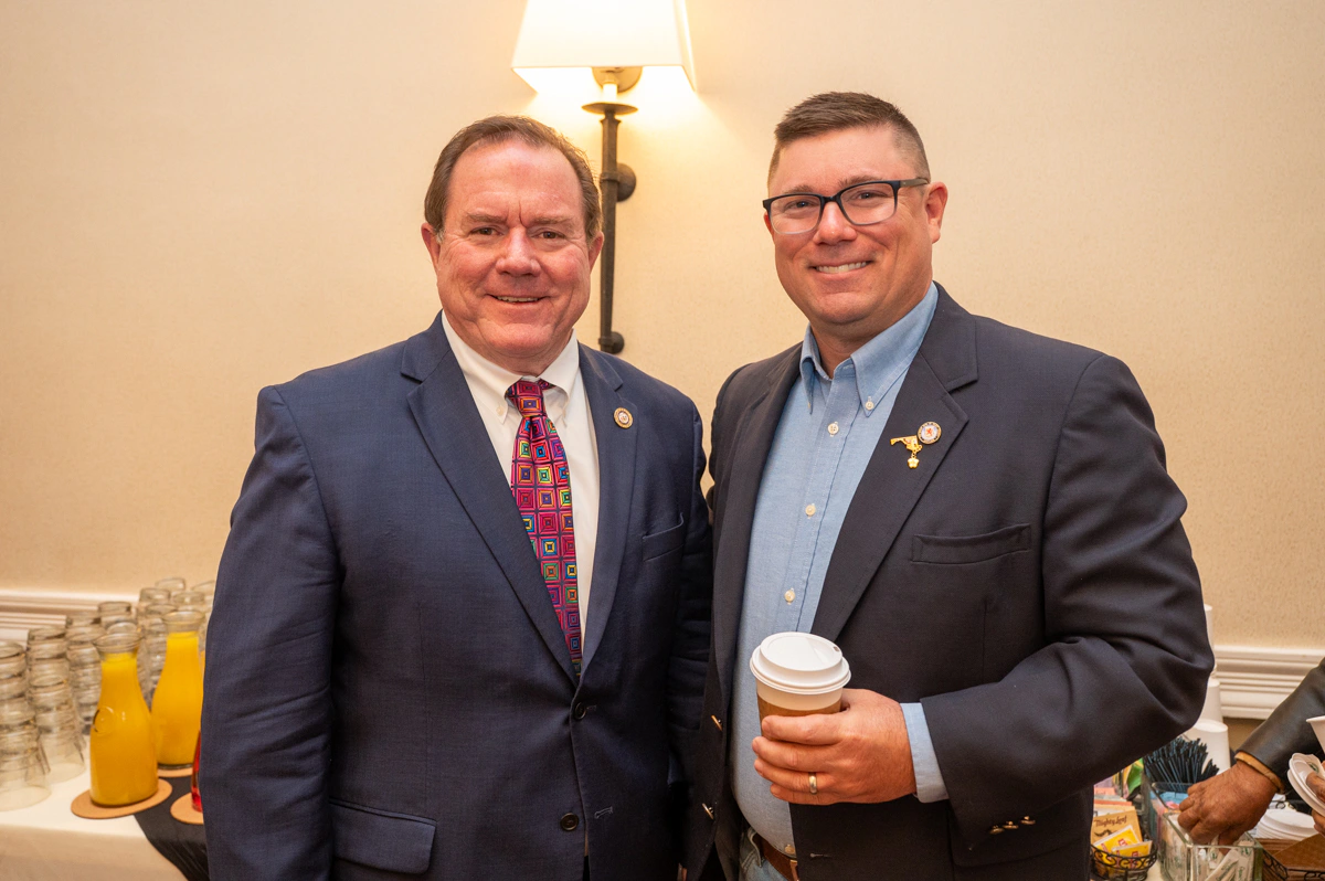Two men in suits stand together, the man on the right holding a coffee cup