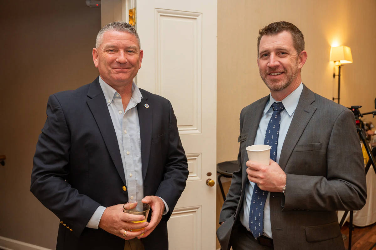 Two men in suits stand together indoors, one holding a coffee cup