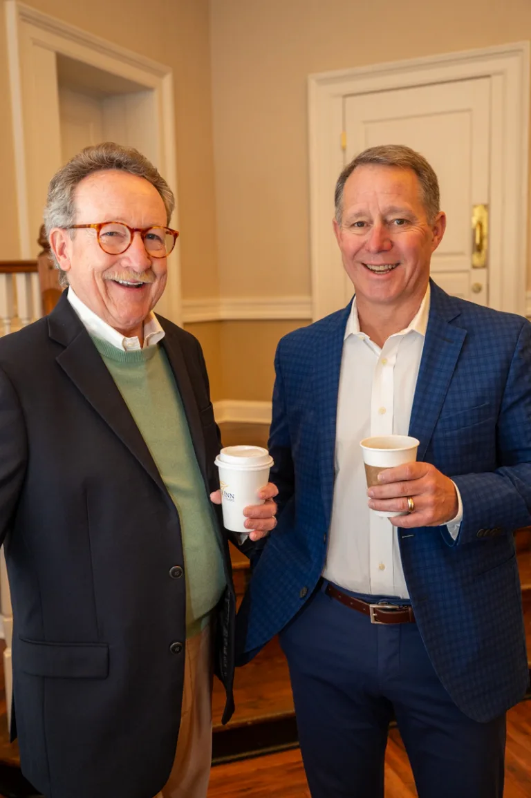 Two older men smile holding coffee cups, one in a dark jacket and one in a navy blazer
