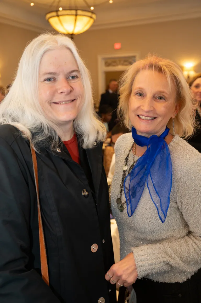 Two smiling women with white hair pose together, one wearing a blue scarf