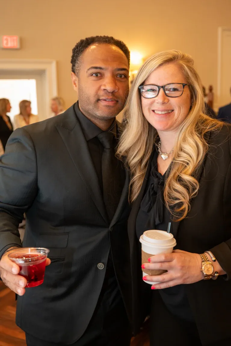 Man in black suit and woman with glasses hold drinks and smile together