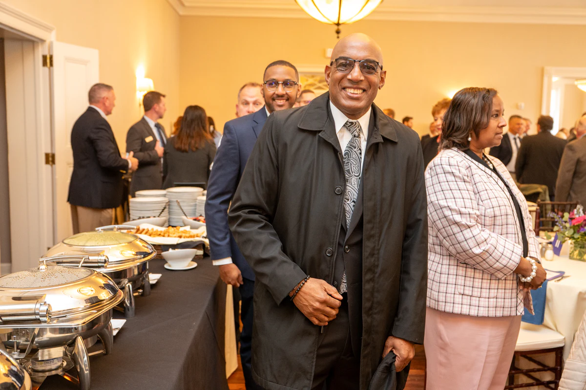 Man in dark coat and gray tie smiles beside a woman in plaid jacket, with food stations nearby