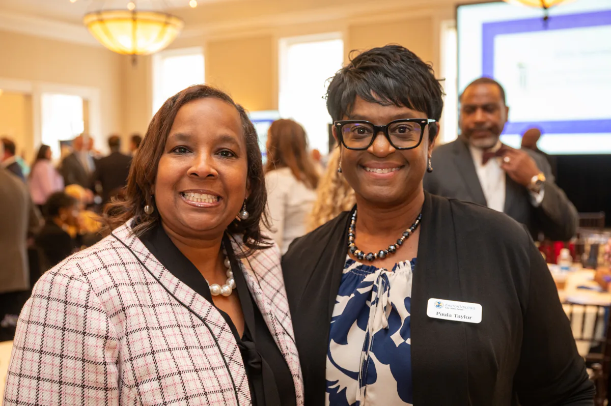 Two women smile at an event, one in a plaid jacket and one with glasses in a blue and white dress