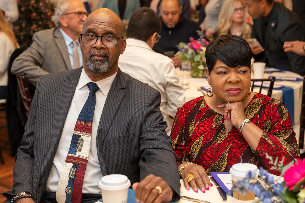 Older man in gray suit and striped tie and woman in patterned red dress sit at a banquet table