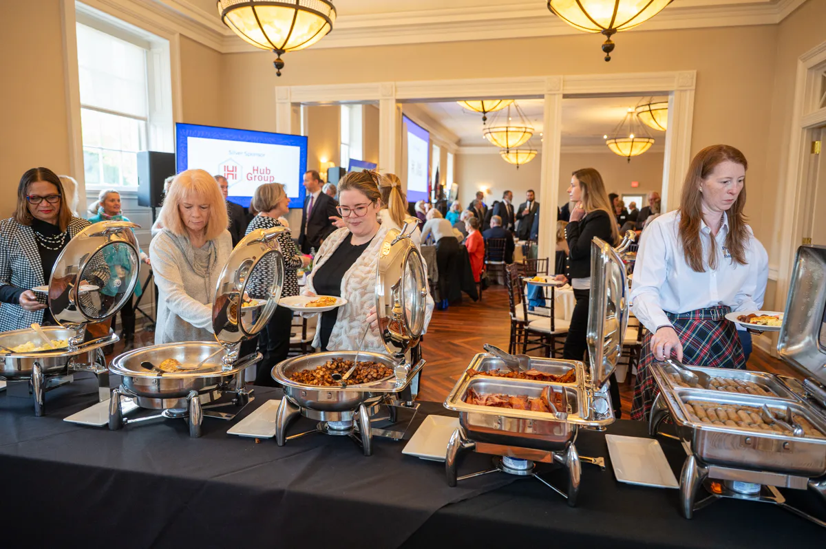 Staff serve food from chafing dishes at a buffet table in a banquet room