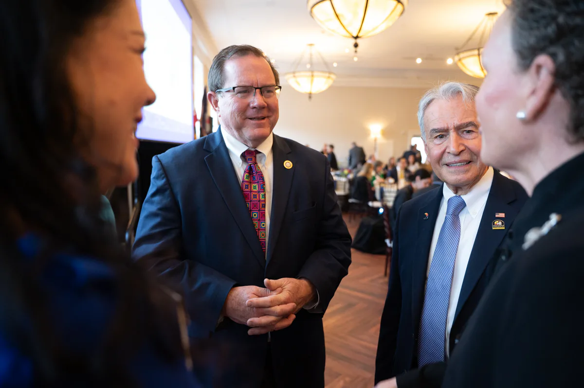 Men in suits talk with a woman in the foreground at a formal reception