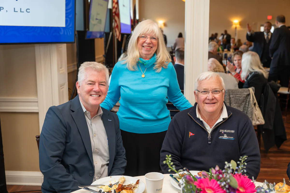 Woman in aqua sweater stands behind two smiling men seated at a banquet table