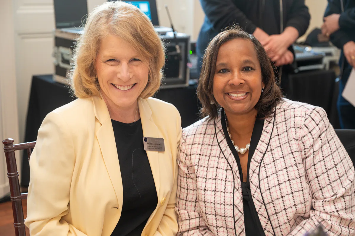 Two women smile side by side, one in a yellow blazer and one in a plaid jacket