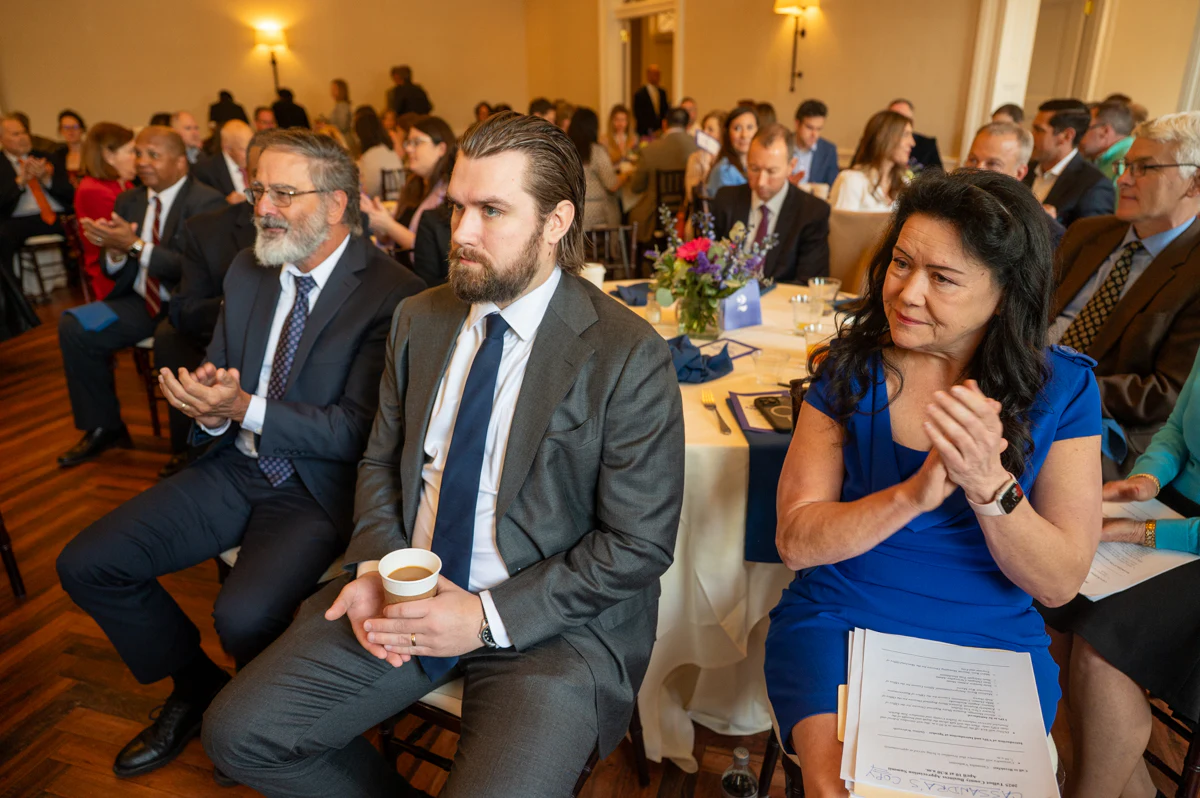 Bearded man and man in gray suit sit at an event, with a woman in blue beside them