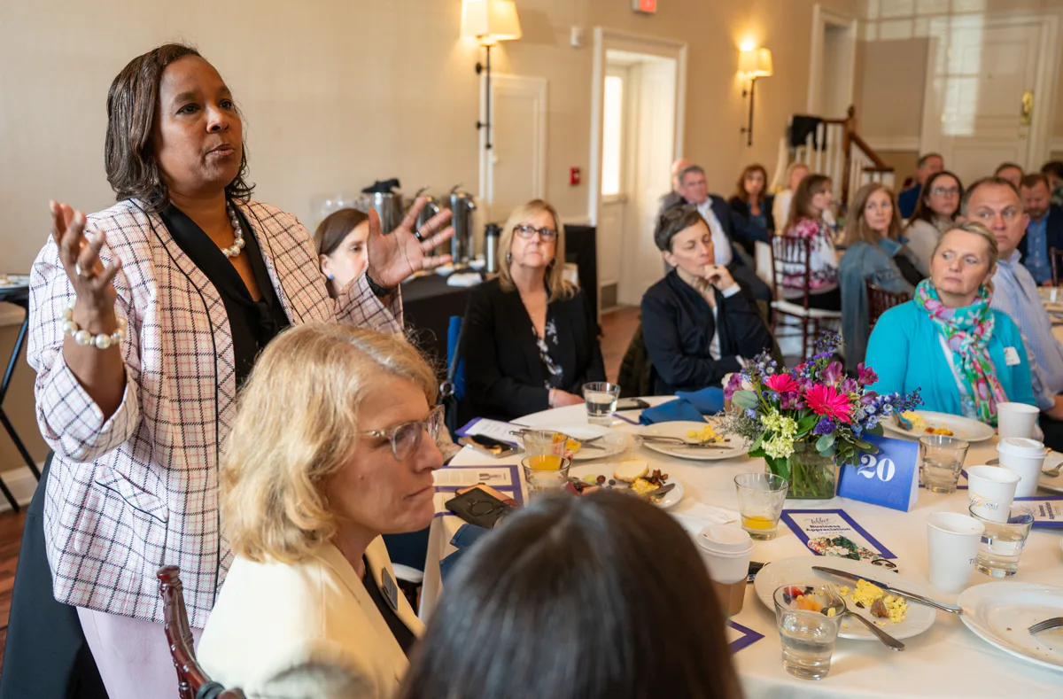 Woman in plaid jacket stands and gestures while speaking at a banquet table