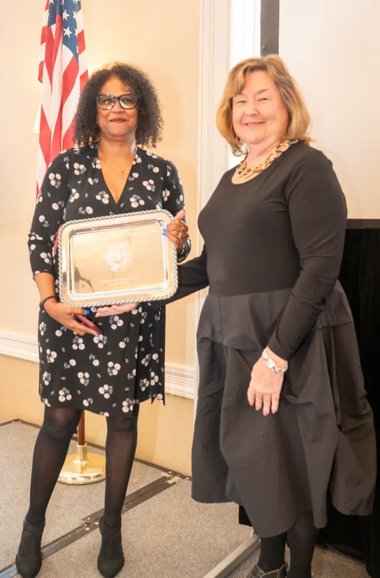 Woman in black floral dress holds a silver tray beside a woman in black, with an American flag behind