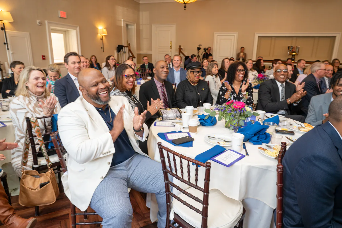 Man in white jacket applauds while seated at a banquet table with other guests