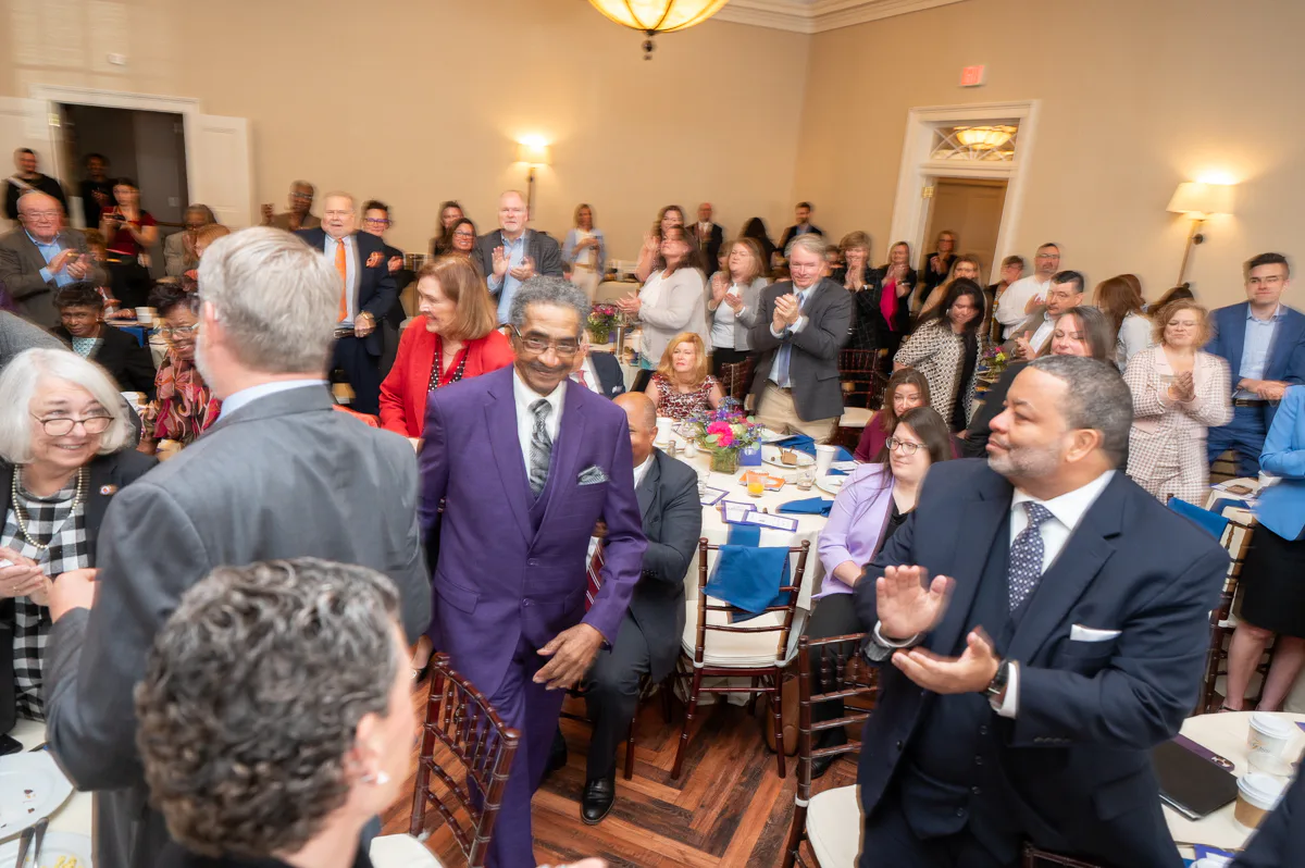 Man in purple suit walks through a crowd of applauding guests