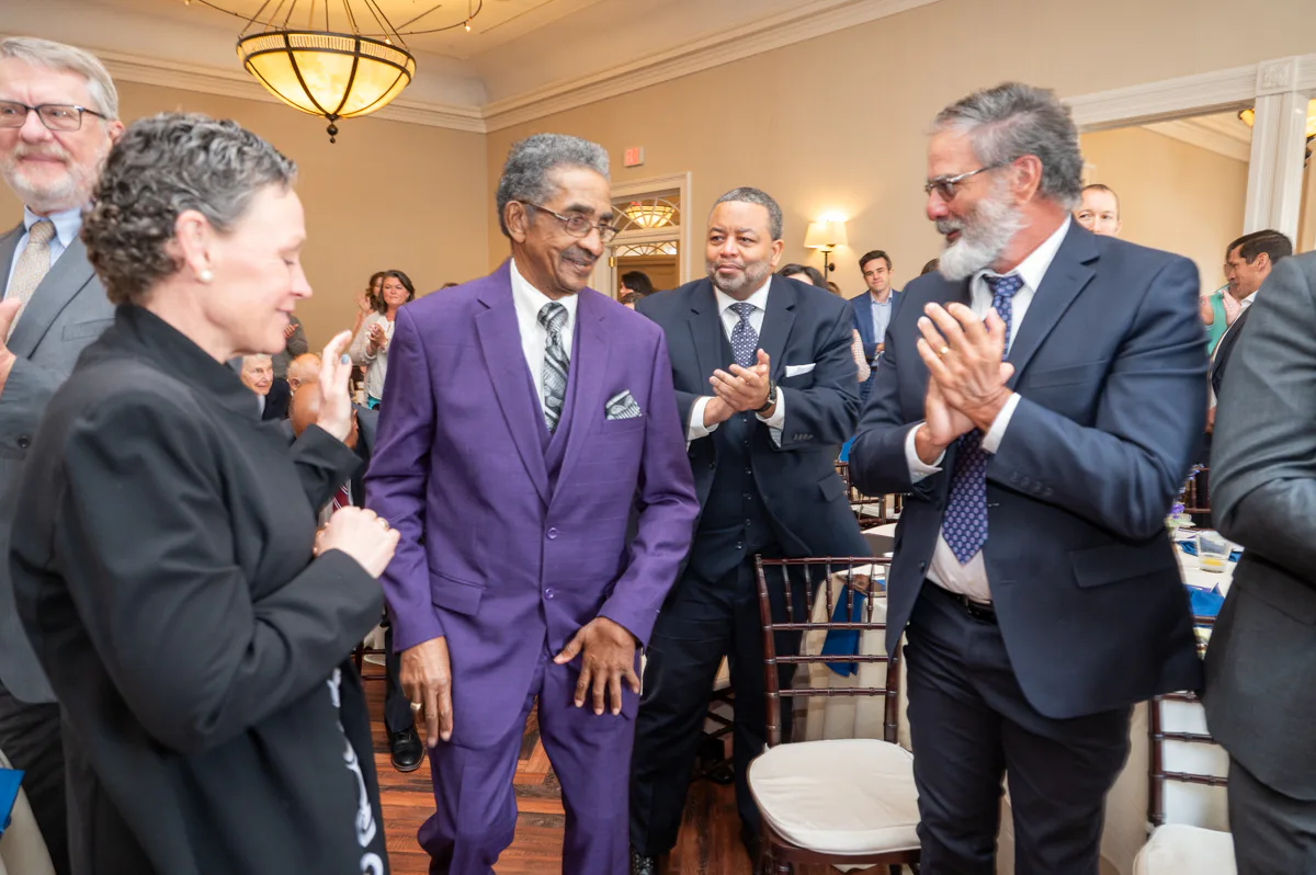 Man in purple suit walks among applauding guests at a banquet