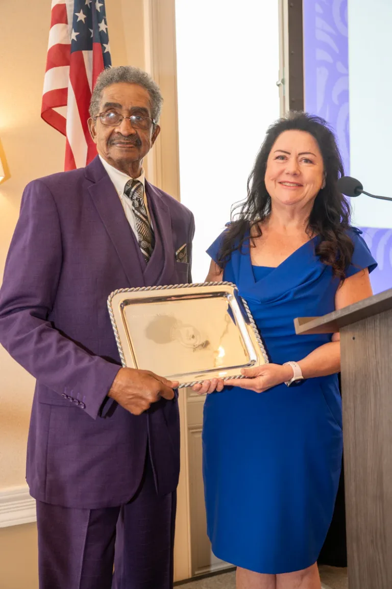 Man in purple suit and woman in blue dress hold a silver tray in front of an American flag