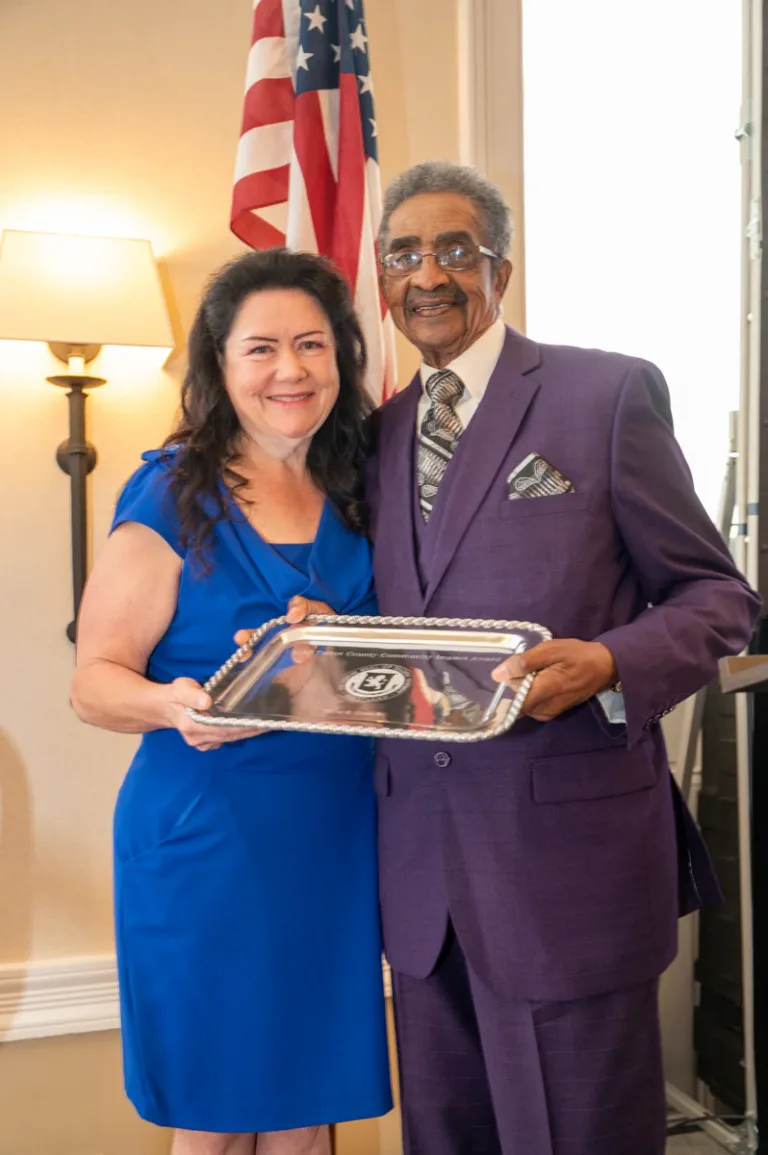 Woman in blue dress and man in purple suit hold a silver tray in front of an American flag