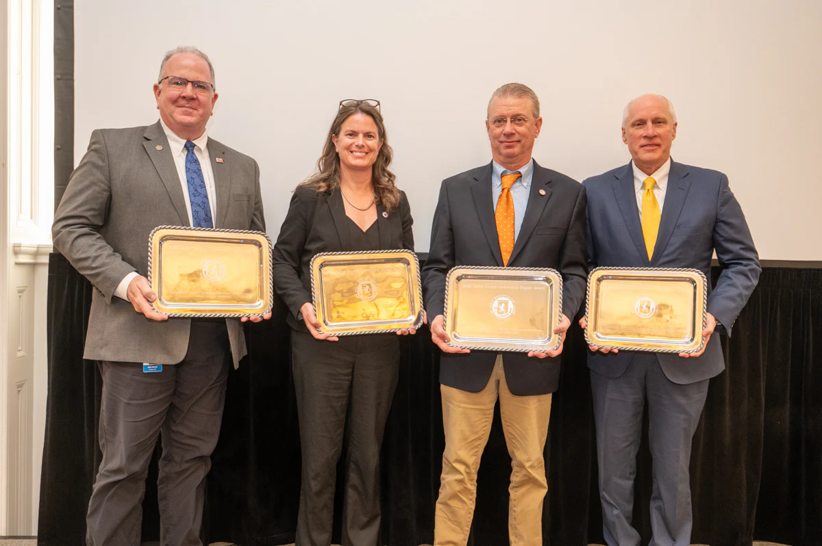 Four award recipients holding gold plaques at an event in a banquet room