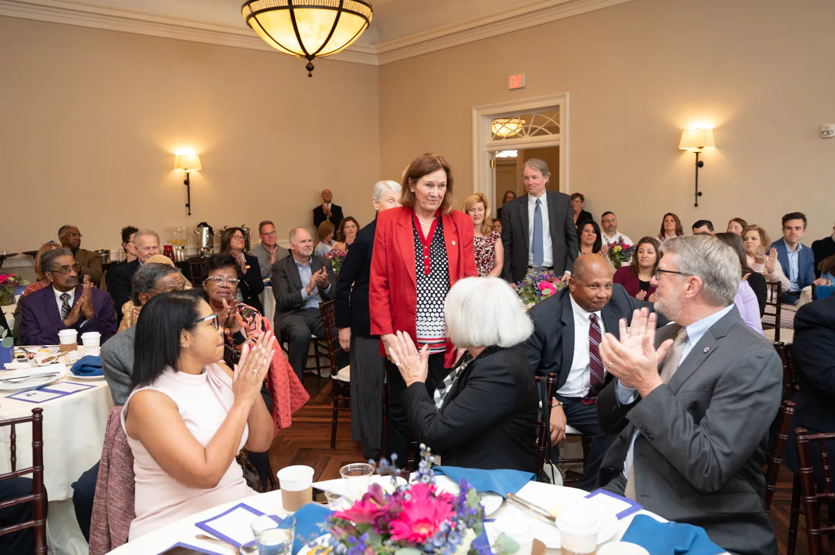 Woman in red jacket standing among applauding guests at a banquet
