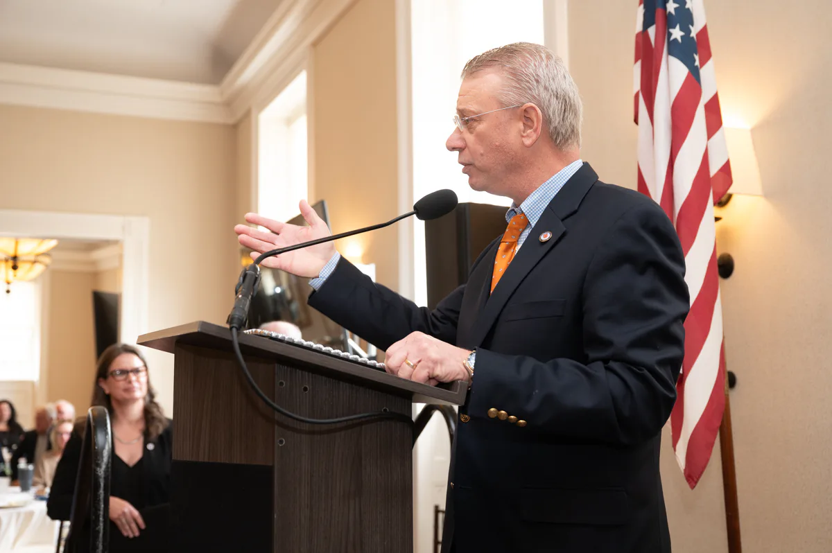 Man in navy suit speaking at a lectern beside a U.S. flag