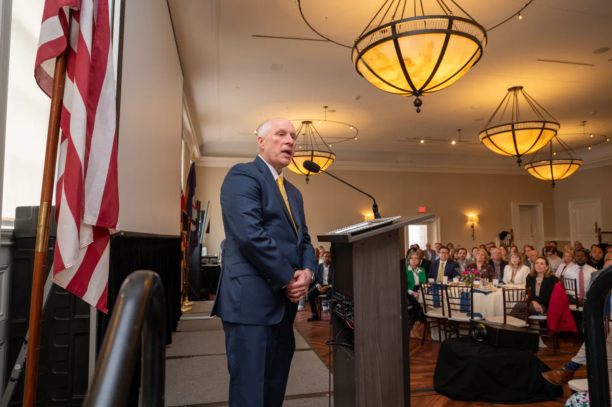 Man in navy suit speaking at a lectern with American and Maryland flags