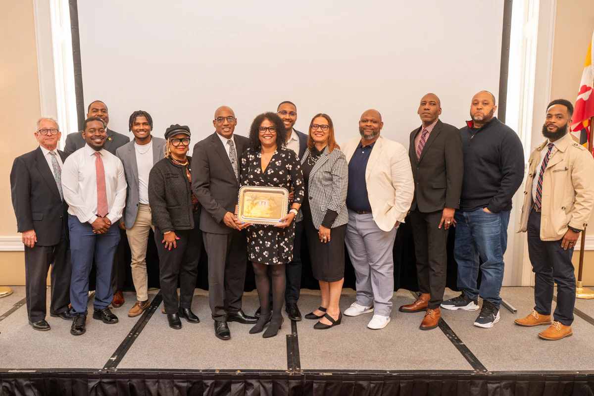Group of about a dozen people posing with a framed certificate in front of a gray wall