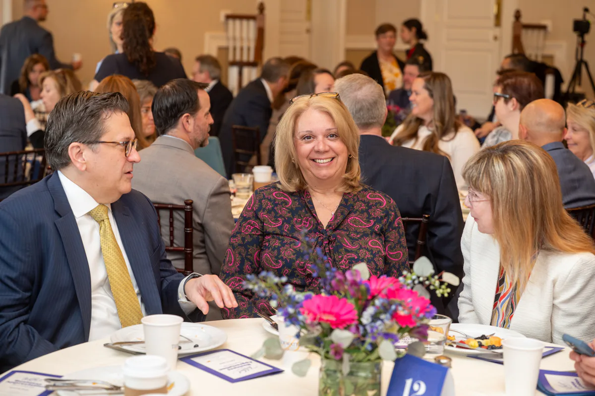 Two people talk over a luncheon table while a woman in a floral top smiles between them