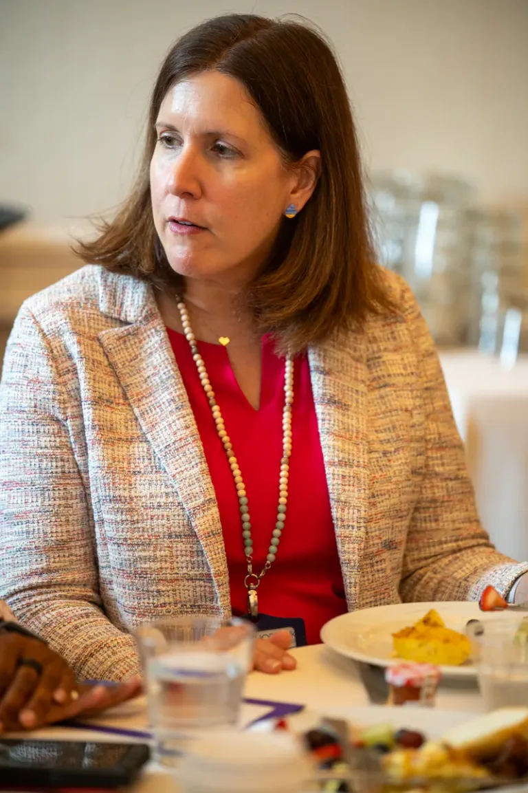 Woman with shoulder-length hair in tweed blazer and red top speaks at a seated event table