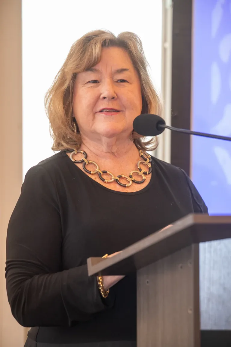 Woman with short gray-blonde hair and beaded necklace speaks at a podium
