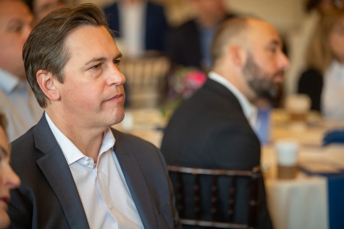 Man in dark suit looks to the side at a seated event, with a bearded man behind him