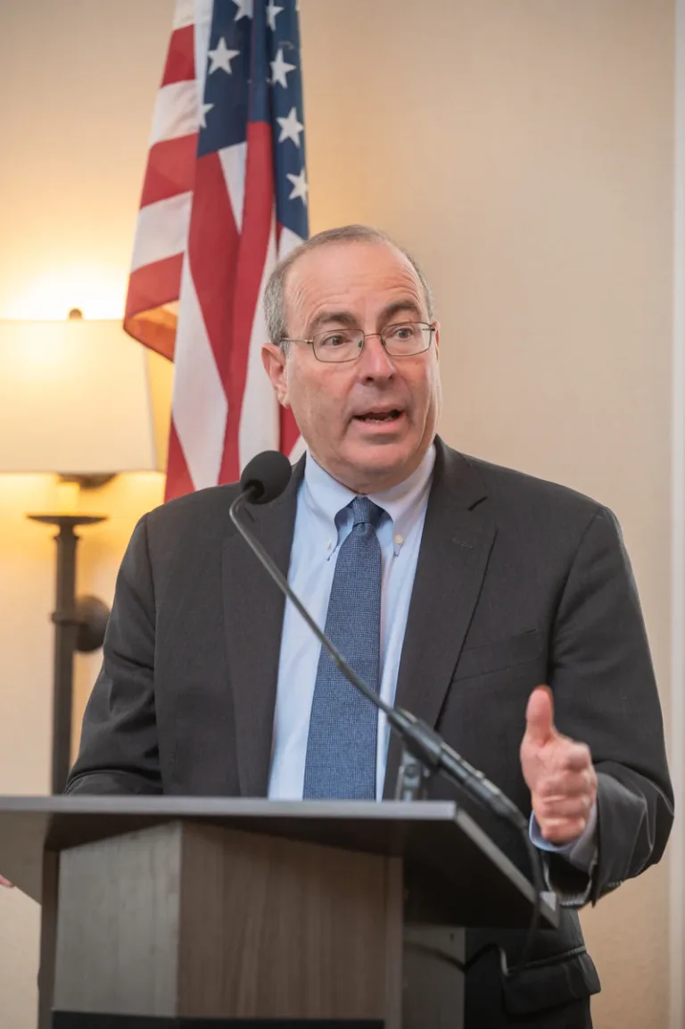 Man in gray suit and glasses speaks at a podium in front of an American flag