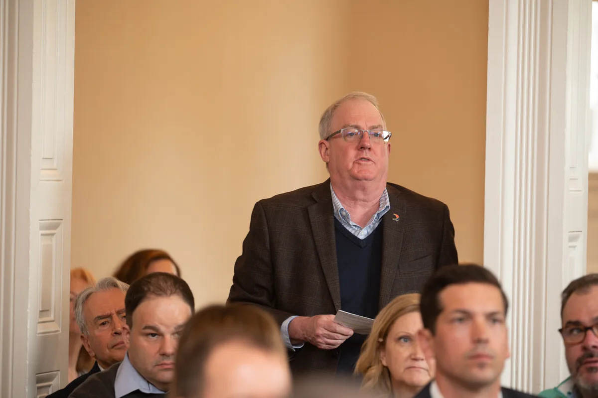 Man in gray sport coat and glasses stands among seated audience members, speaking