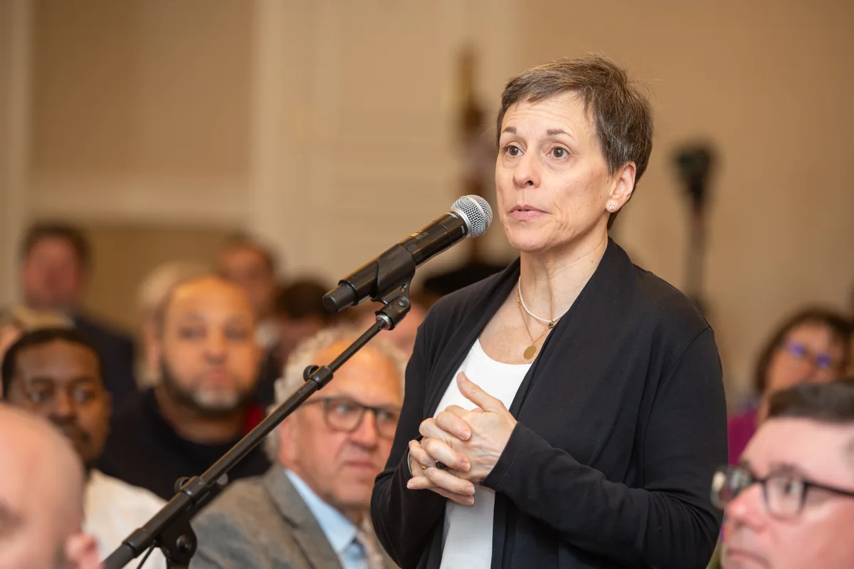A woman with short hair speaks into a microphone to a seated audience.