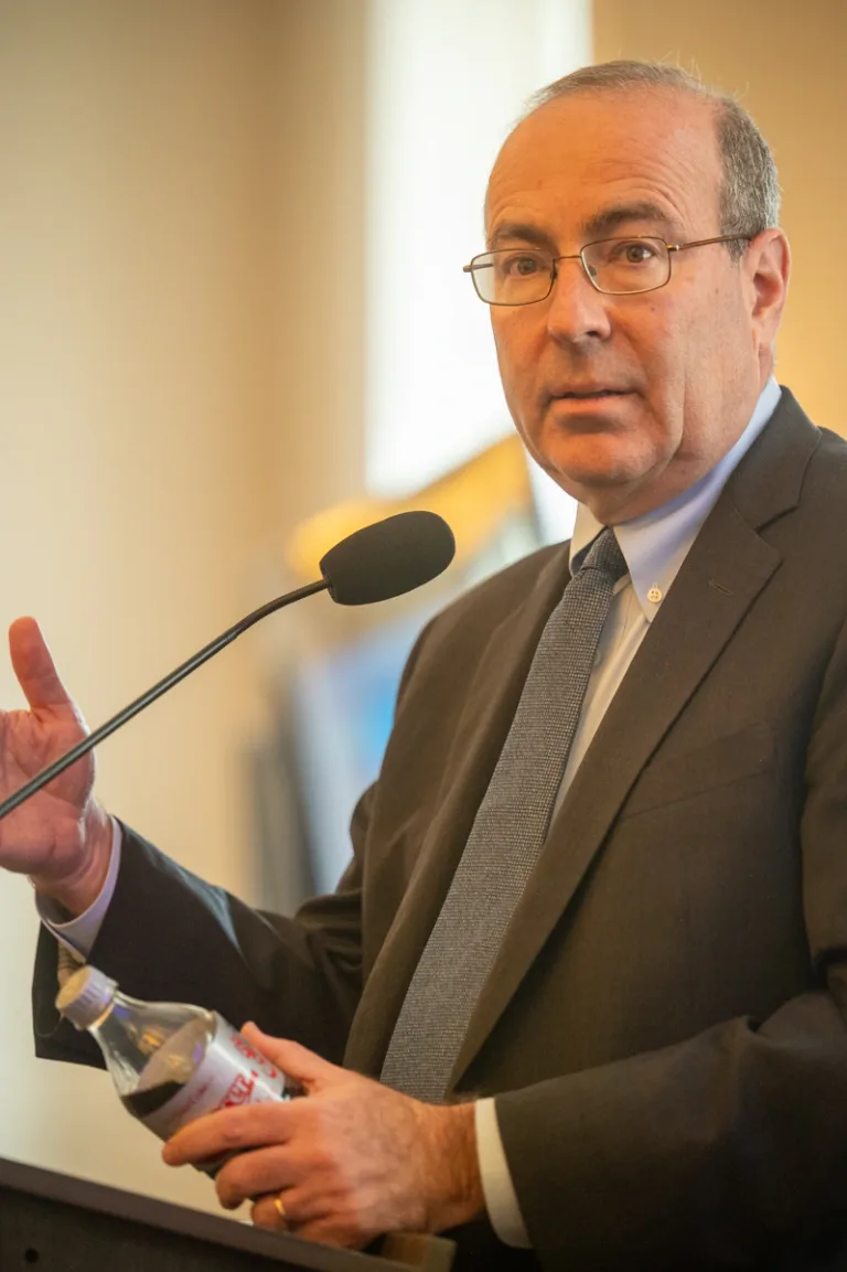 Man in gray suit and glasses gestures while speaking at a wooden podium