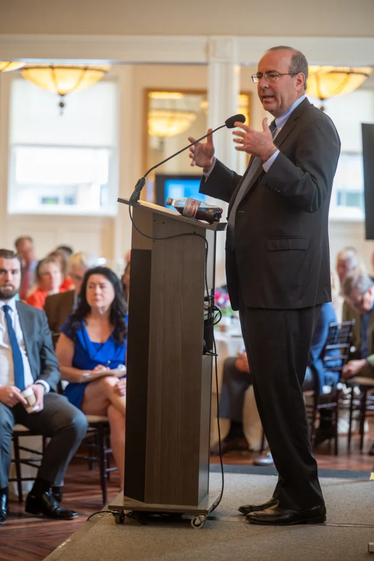 Man in dark suit speaks at a podium and gestures with one hand in a formal room