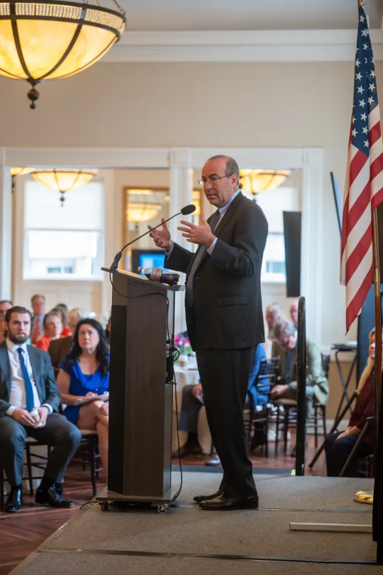 Man in dark suit speaks at a podium under a chandelier, with an American flag beside him