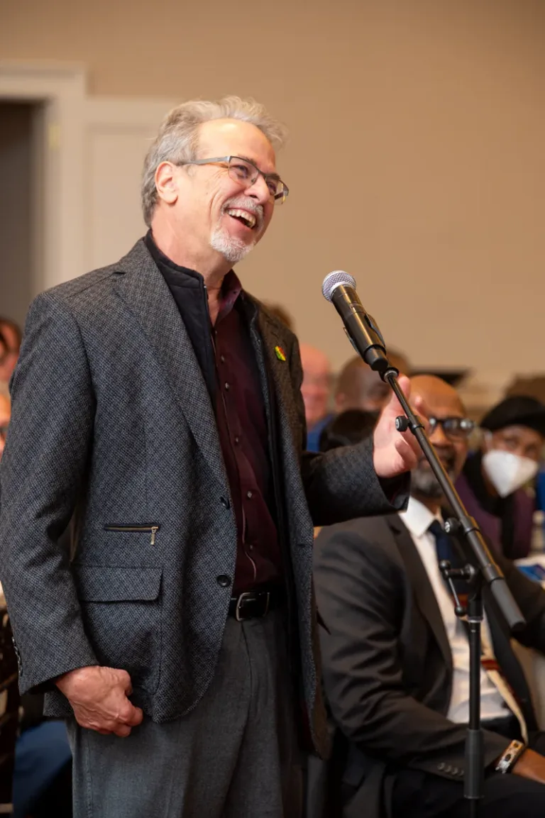 Man in gray sport coat and glasses laughs while speaking into a microphone