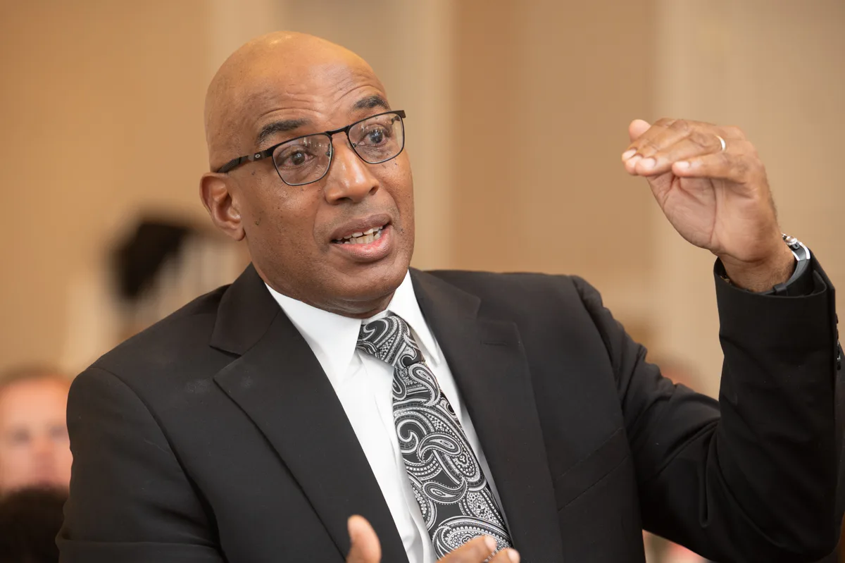 Bald man with glasses and patterned tie gestures while speaking