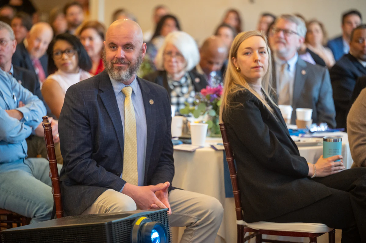 Bearded man in blue blazer and blonde woman sit at an event with an audience behind them