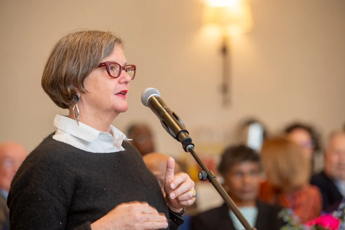 Woman with short hair and red glasses speaks into a microphone at an event