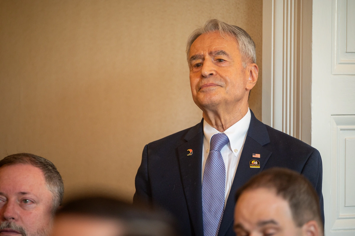Gray-haired man in navy suit with lapel pins stands and listens in a room