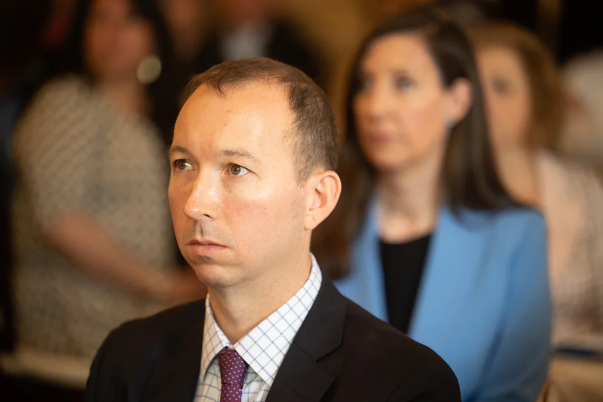 Man in dark suit listens intently at an event, with a woman in a blue blazer behind him