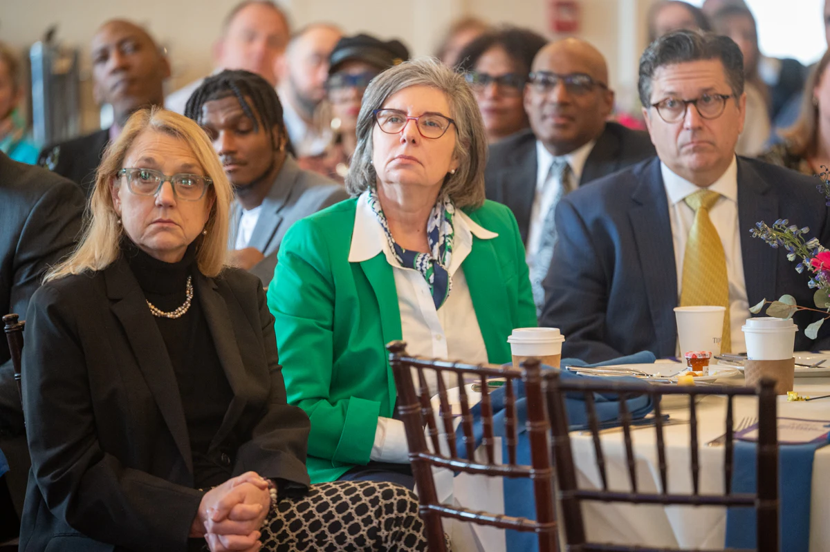Woman with glasses and green blazer sits beside a man in a suit at an audience table